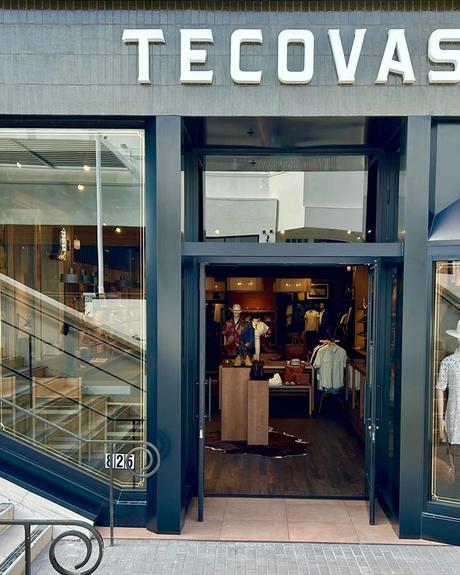 Exterior view of a Tecovas store with glass doors and windows displaying boots and clothing. Stairs are on the left side, and a neon "BOOTS" sign is visible inside.