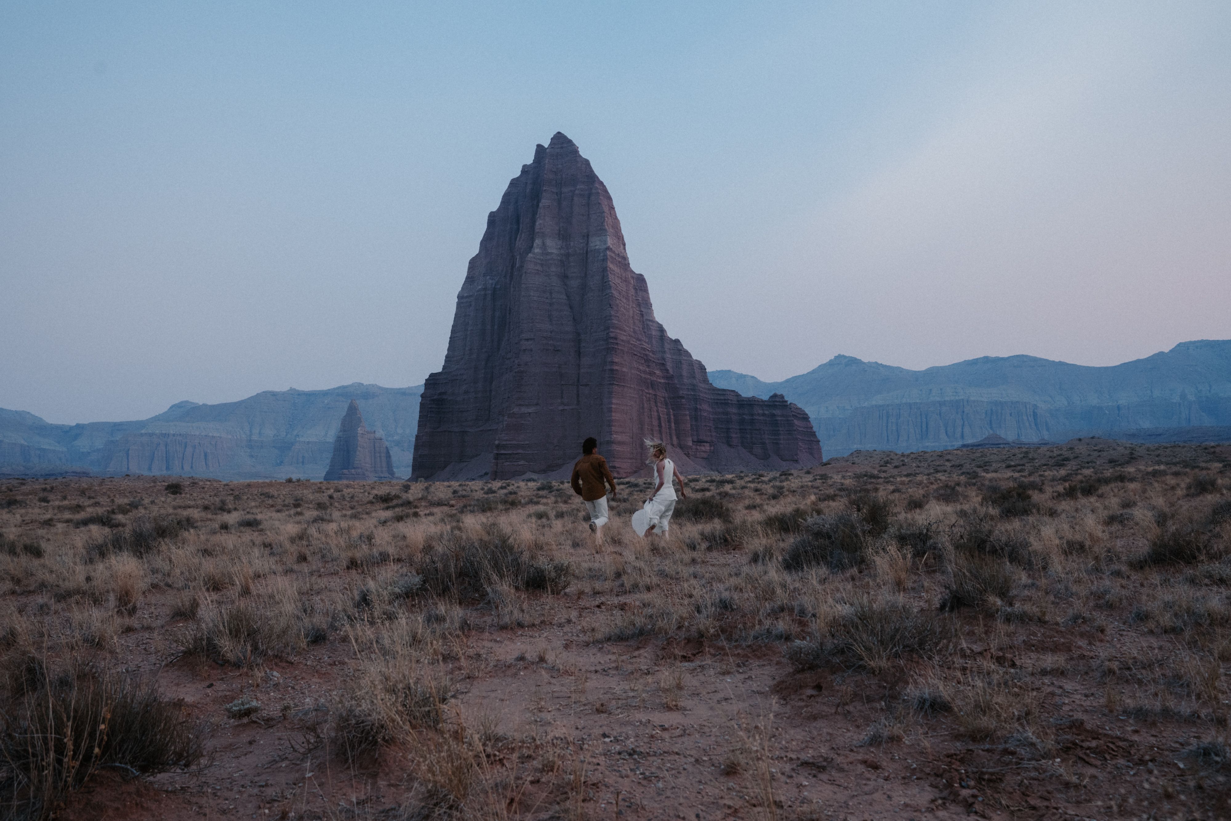 A couple walks through a desert landscape toward a large, pointed rock formation.