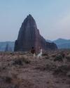 A couple walks through a desert landscape toward a large, pointed rock formation.