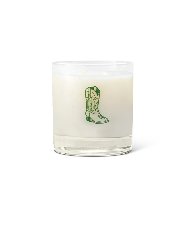 A white candle in a clear glass holder with a green illustration of a cowboy boot on the front.
