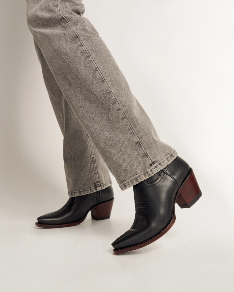 Person wearing gray jeans and black leather heeled cowboy boots with brown soles, standing on a white background.
