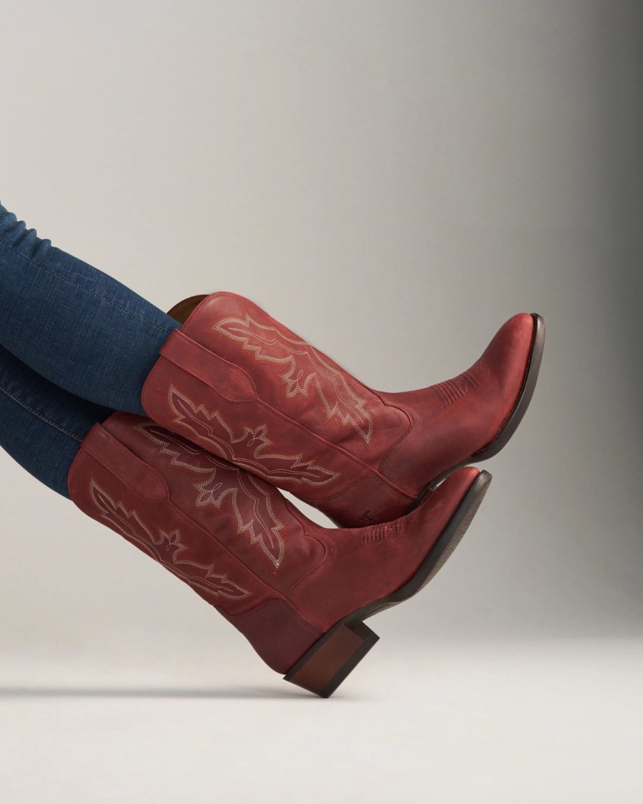 The Britt in Redwood Goat. A person wearing dark blue jeans and striking redwood leather cowboy boots with decorative stitching, feet raised against a neutral background.