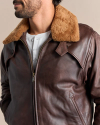 Close-up of a person wearing a dark brown leather jacket with a beige shearling collar, over a white buttoned shirt. The person has a beard and is looking to the side.