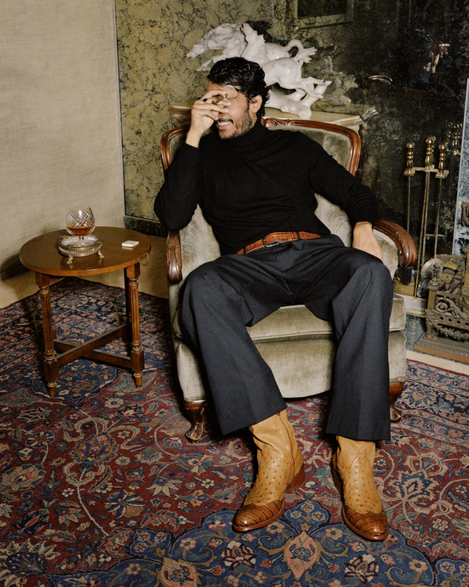 A man in a black turtleneck and dark trousers sits in an armchair, holding a cigar to his face, wearing tan textured boots.
