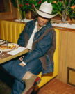 Person in a cowboy hat and denim outfit with fringe details sits in a restaurant booth beside a table with food.