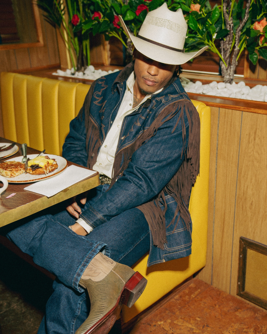 Person in a cowboy hat and denim outfit with fringe details sits in a restaurant booth beside a table with food.