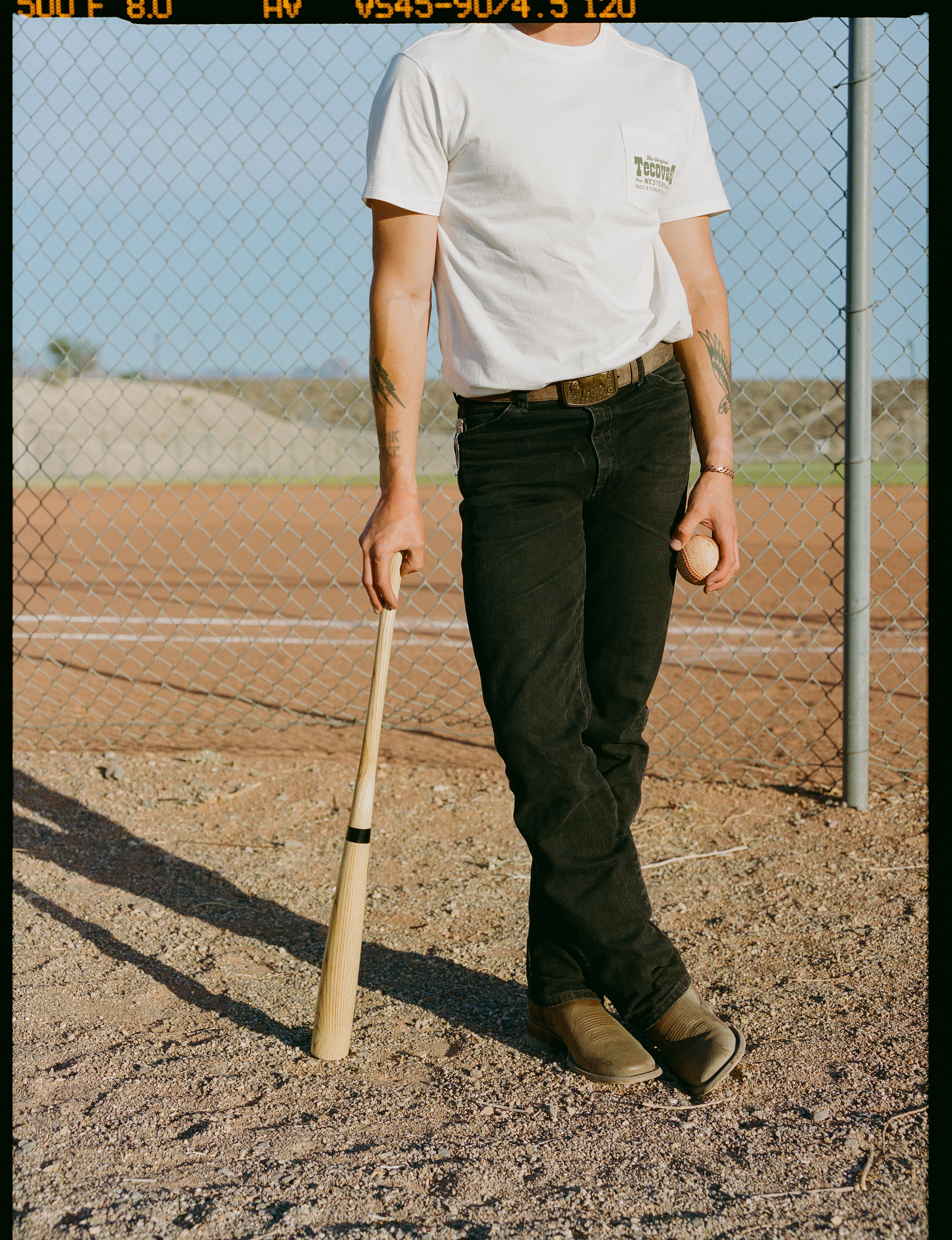 Person in a white t-shirt and black jeans stands on a baseball field holding a baseball bat and a baseball, with a chain link fence in the background.