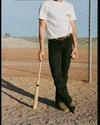Person in a white t-shirt and black jeans stands on a baseball field holding a baseball bat and a baseball, with a chain link fence in the background.