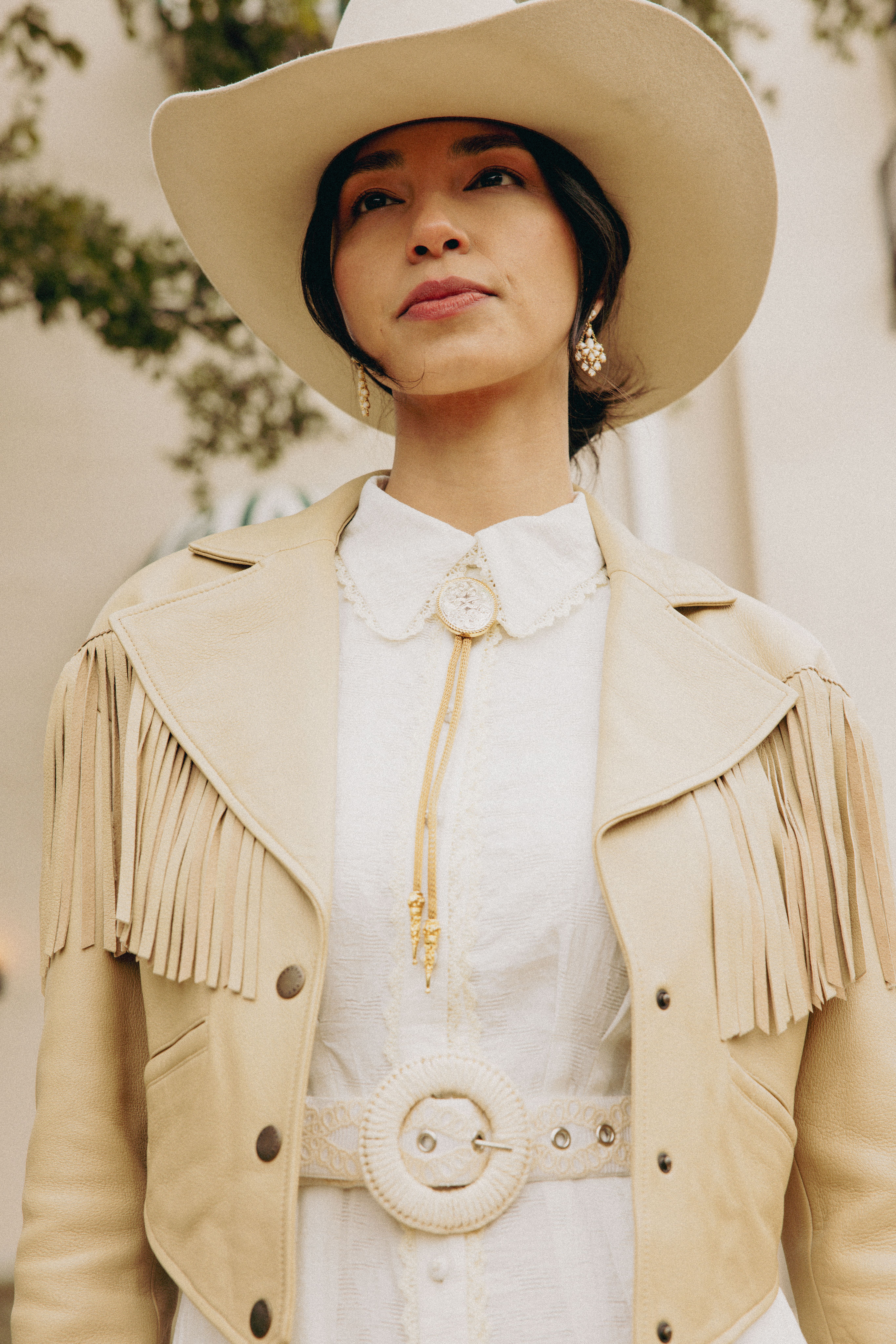 Woman in a white dress and fringed leather jacket wears a wide-brimmed hat outdoors.