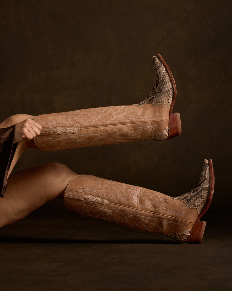 The Loretta in Mangrove Invasive Python. A person lies on their back, holding up their legs wearing The Loretta Invasive Python tan leather cowboy boots with striking snakeskin patterns and wooden heels, against a dark background.