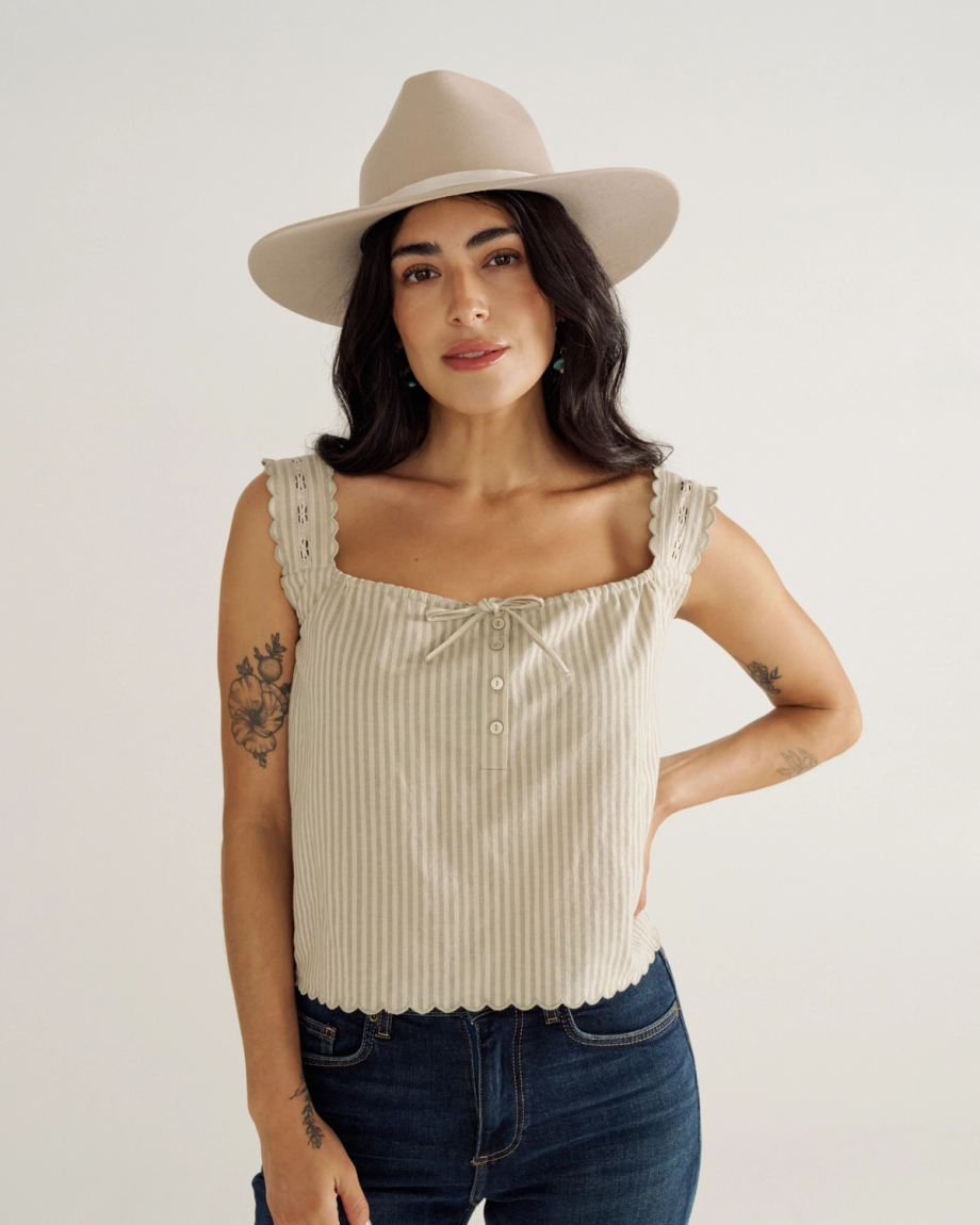 Woman wearing a beige hat and sleeveless ribbed top stands against a plain background with one hand on her hip.