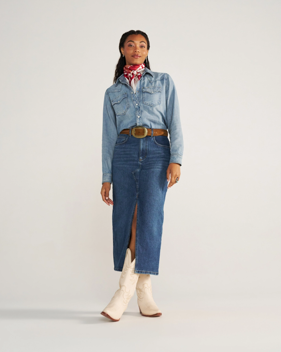 A woman stands wearing a denim shirt, denim skirt with a front slit, white cowboy boots, a brown belt, and a red patterned neck scarf.