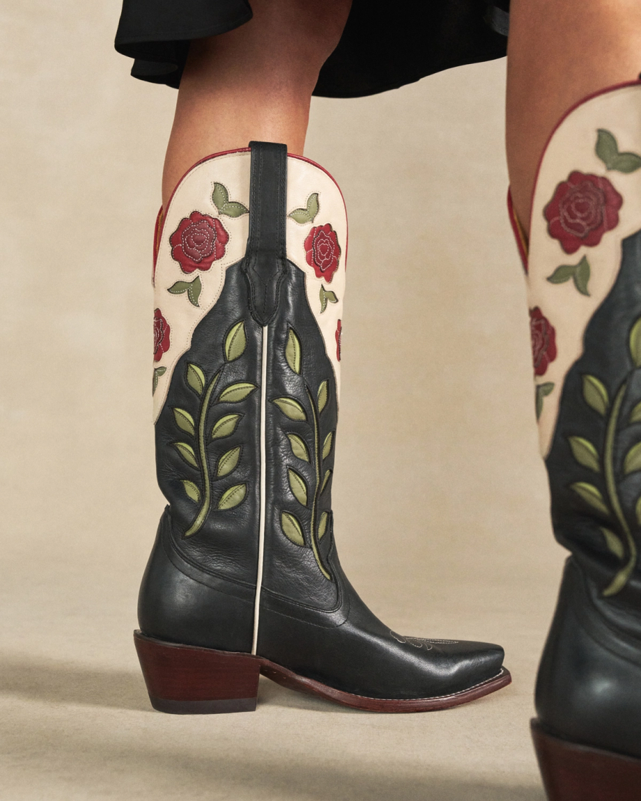 The Annie in Midnight Roses Cowhide. Close-up of a person wearing black Midnight Roses Cowhide cowboy boots with red rose and green leaf embroidery, standing on a neutral background.