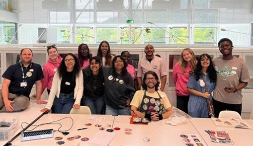 Black Humanities Project Team with Student Ambassadors standing behind Ben from Badge Cafe who is sitting at a large table with a selection of badges on the table.