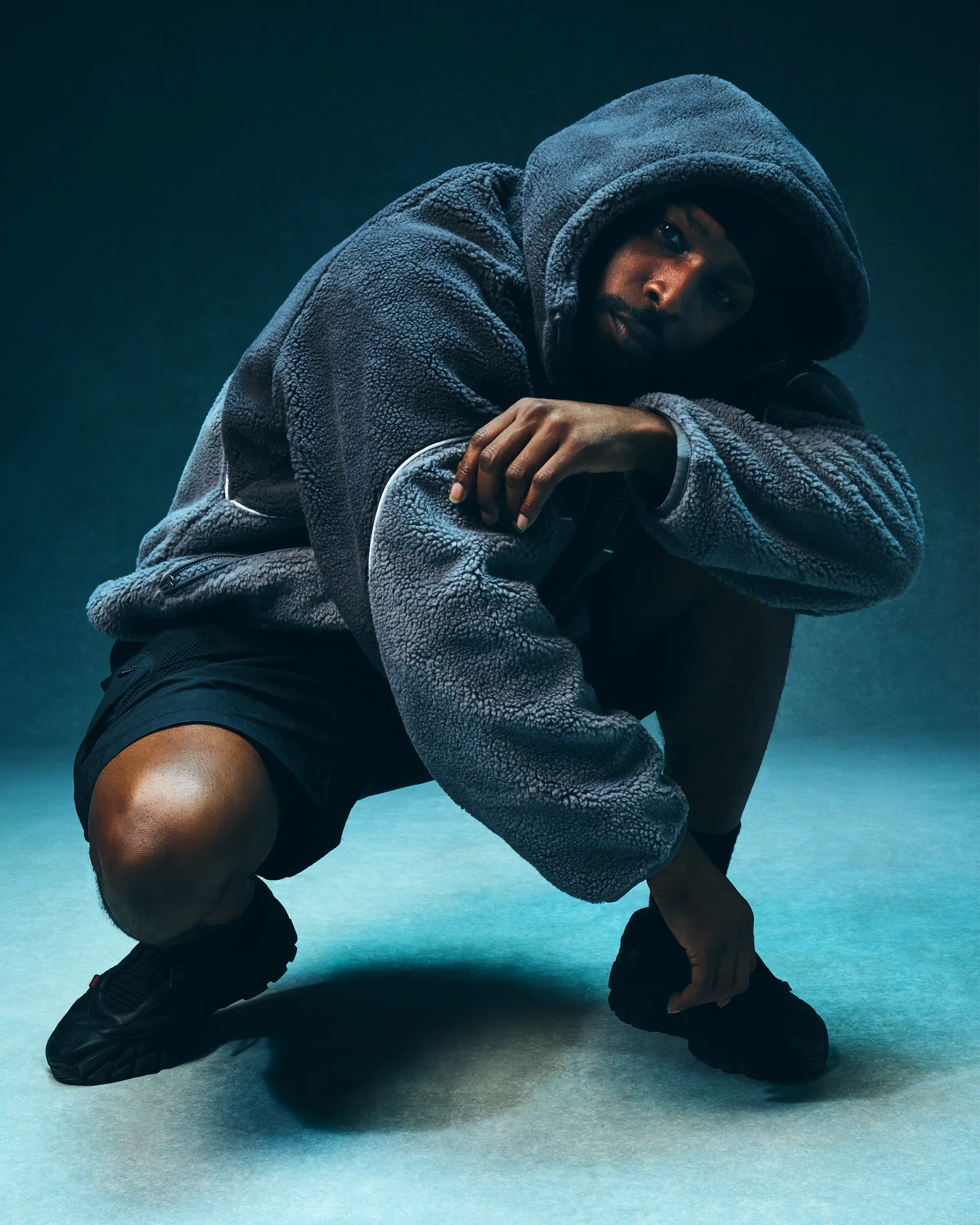 JME in SDMN grey fleece hoodie arms crossed standing wide against teal studio backdrop, full length editorial fashion portrait London