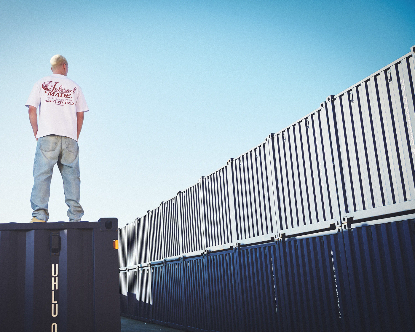 male model in black Internet Made hoodie crouching in front of yellow shipping containers, gold teeth, editorial streetwear campaign photography London