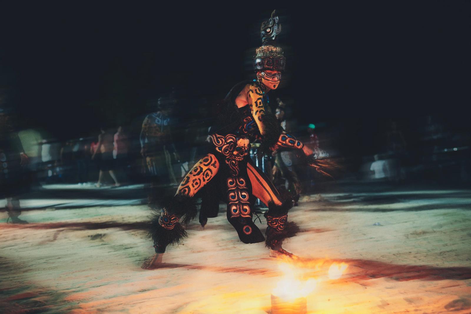 performer in ceremonial body paint crouching beside fire during a contemporary ritual performance on a beach in Playa del Carmen at night