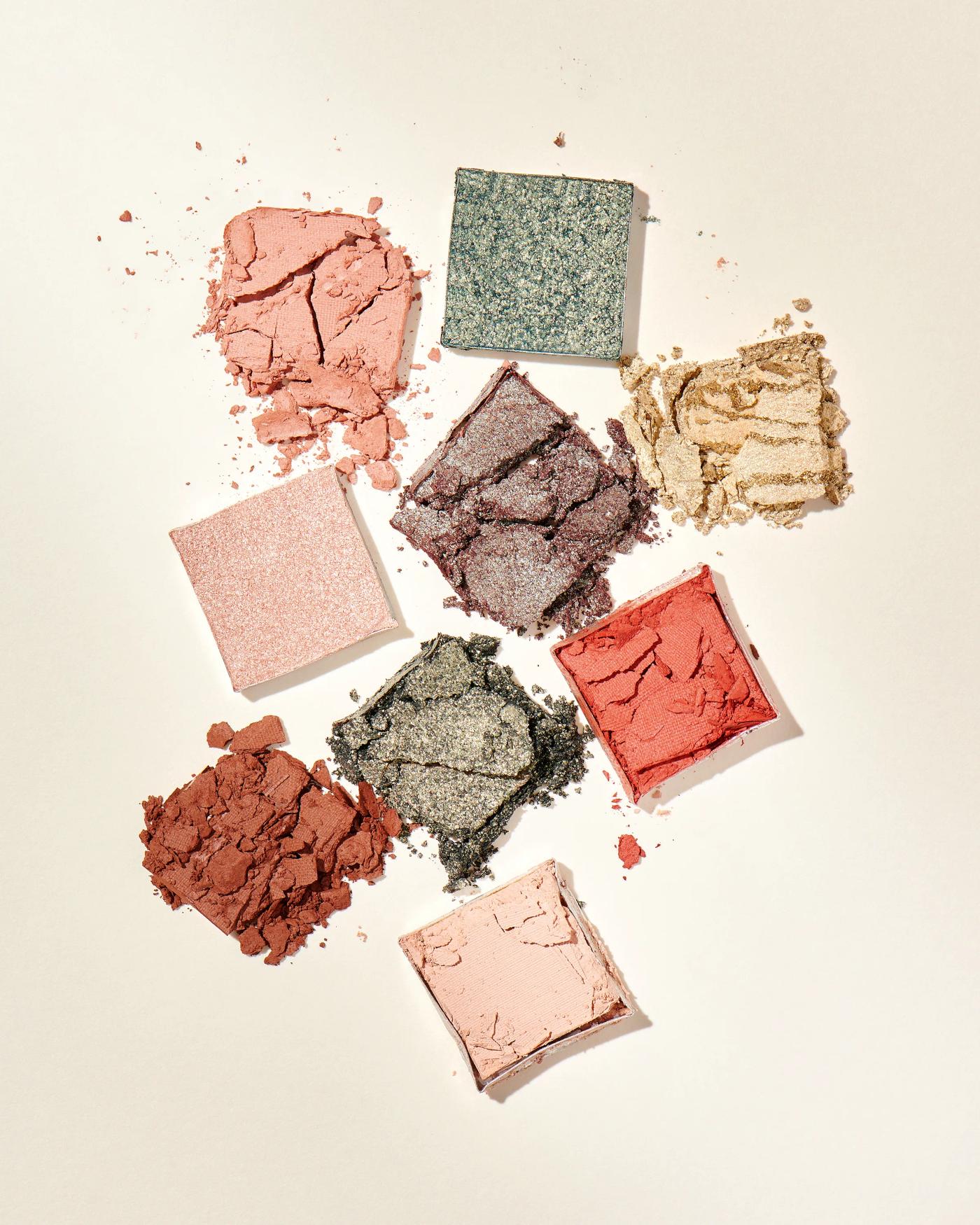 crushed Ciaté London eyeshadow pans in earth and jewel tones scattered on cream surface, beauty photography London