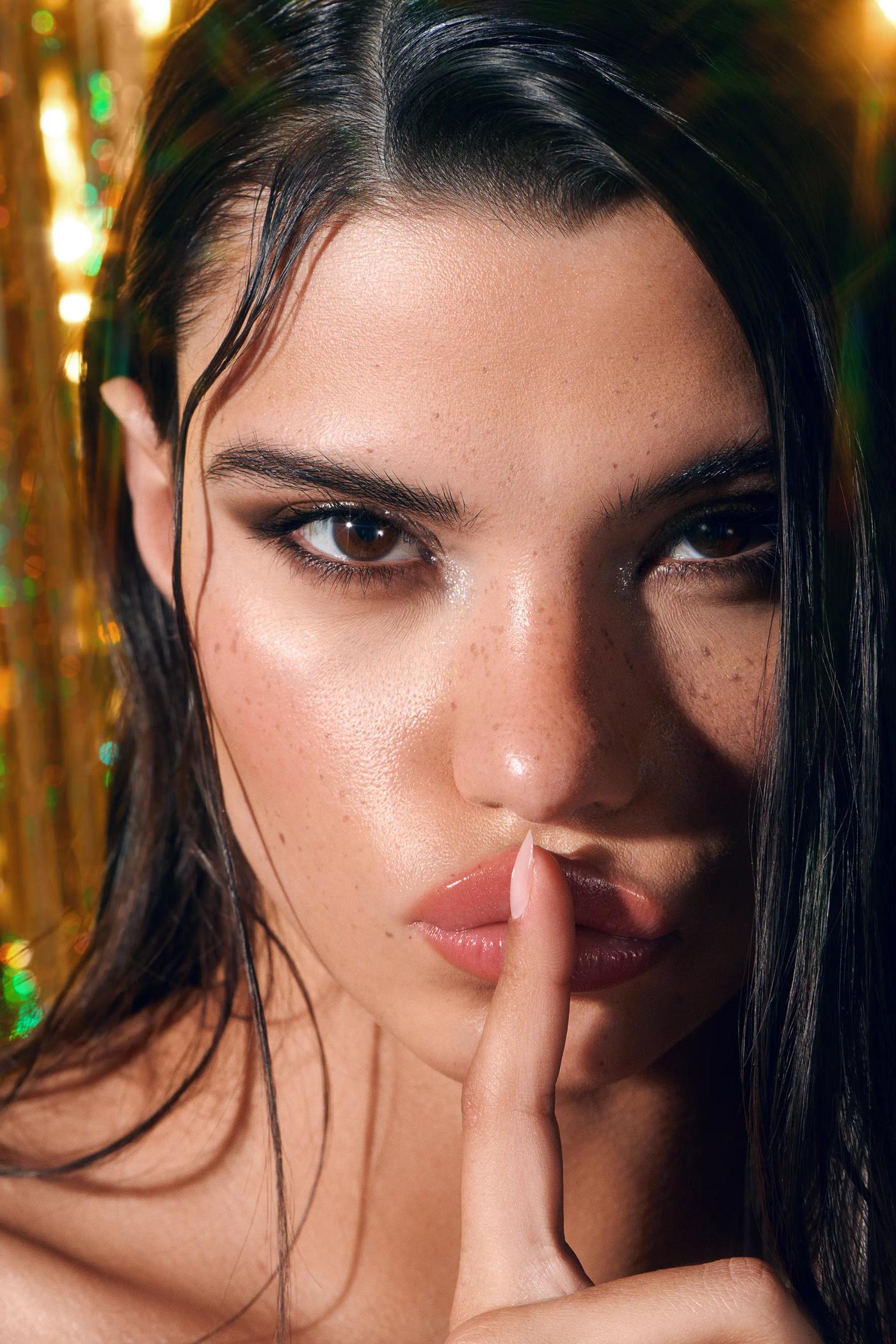 close up portrait of female model with finger to lips, glitter inner corner eye, gold bokeh backdrop, editorial beauty campaign photography London