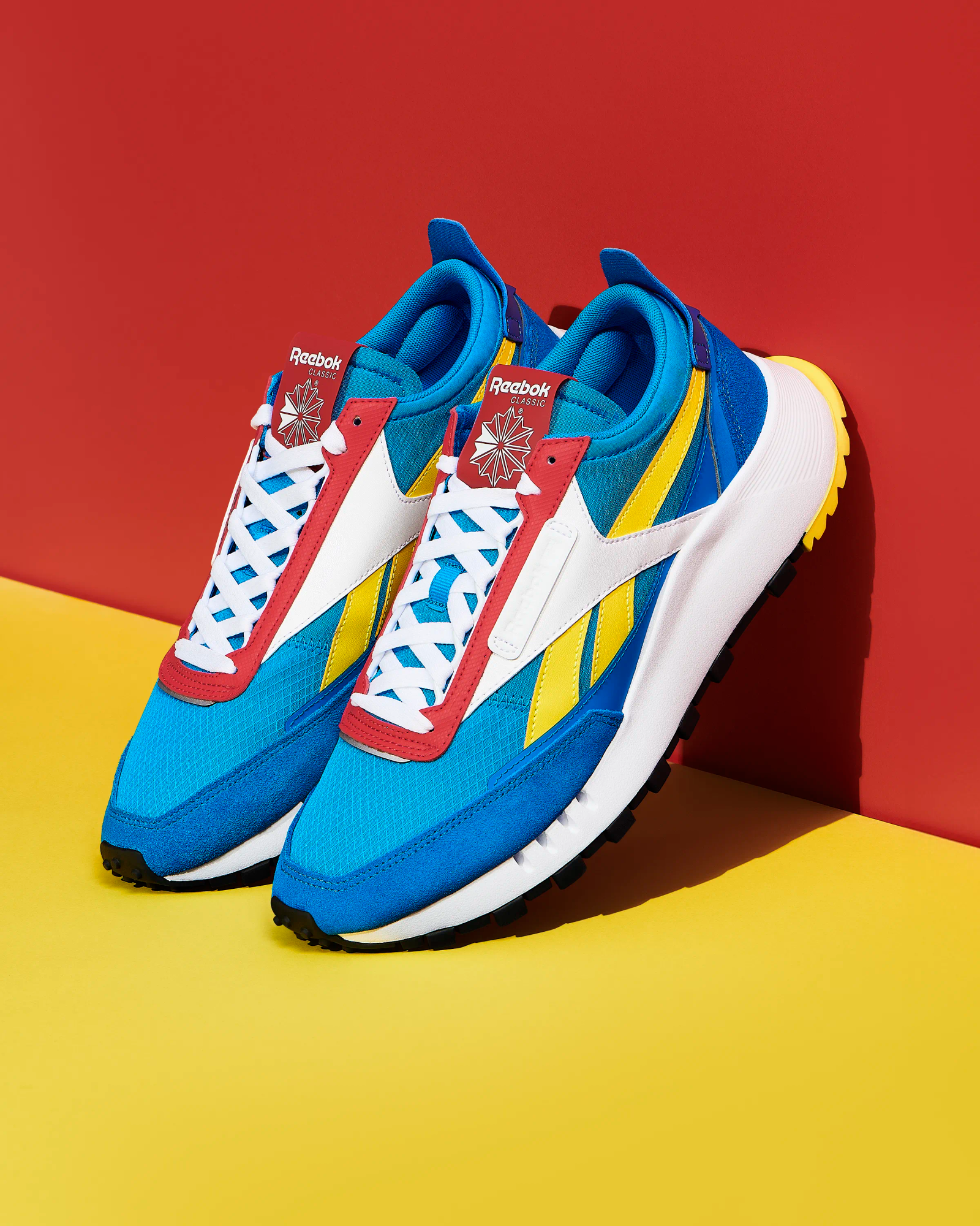 Reebok Classic trainers in blue, yellow and red against bold red and yellow geometric backdrop, footwear photography London