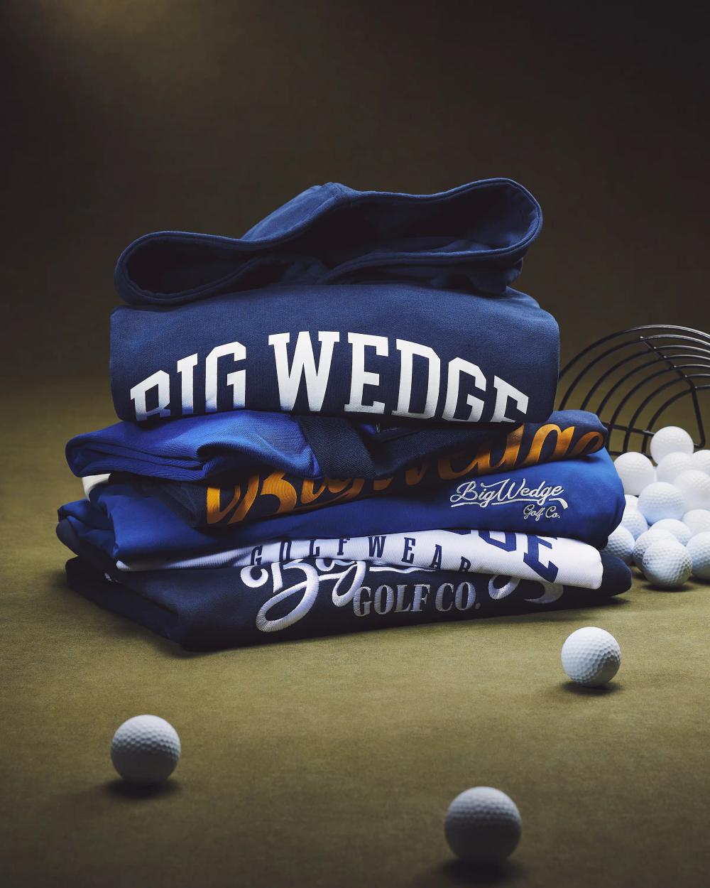 stack of Big Wedge Golf Co. sweatshirts and hoodies in navy with golf balls and wire basket on green surface, sportswear campaign photography London