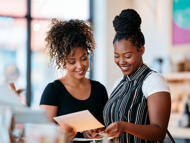A small business owner and team member working together at the front counter of their shop during the day, smiling while going through paperwork.