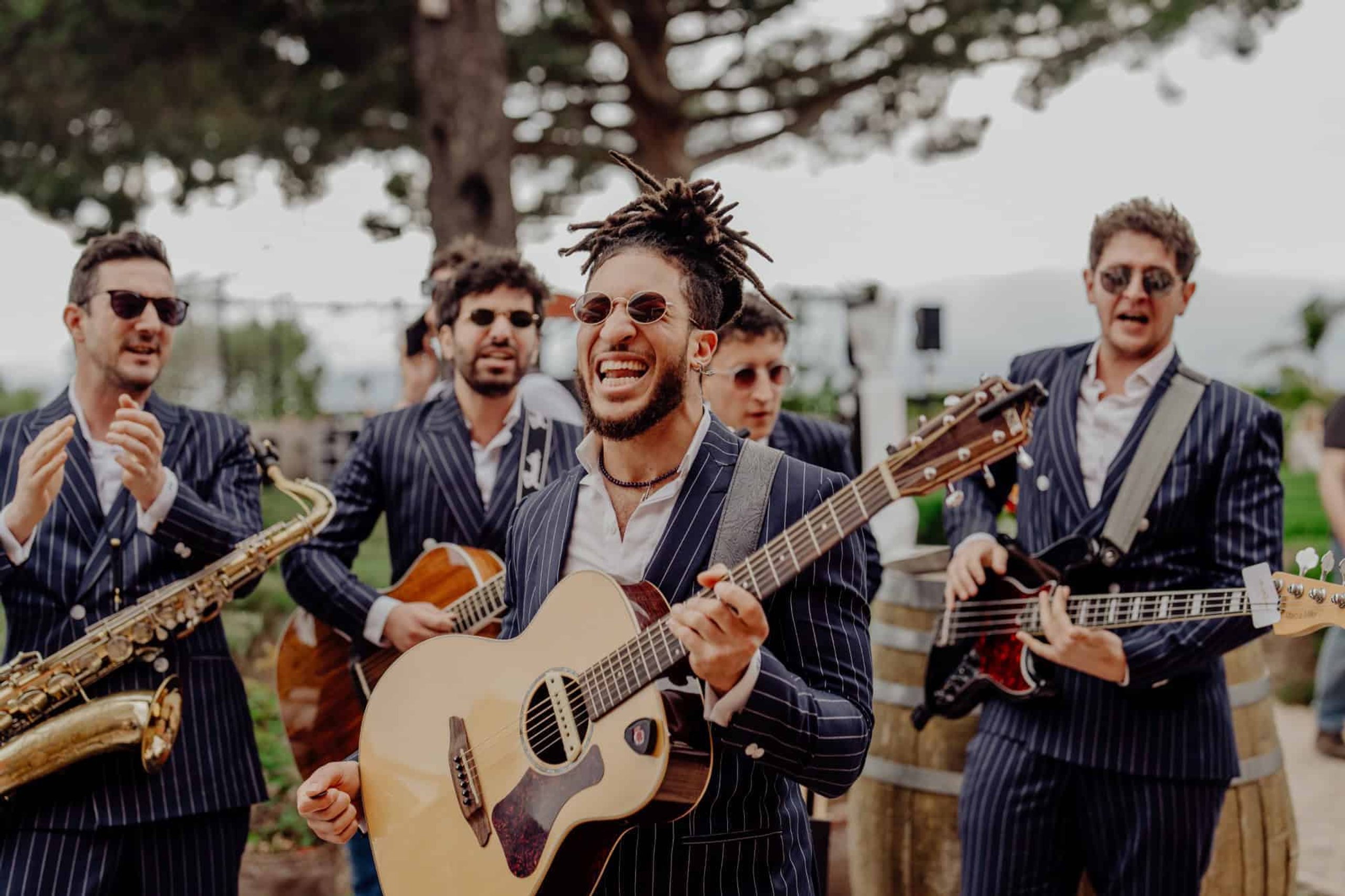 A saxophonist and two guitarists playing outdoors under pine trees