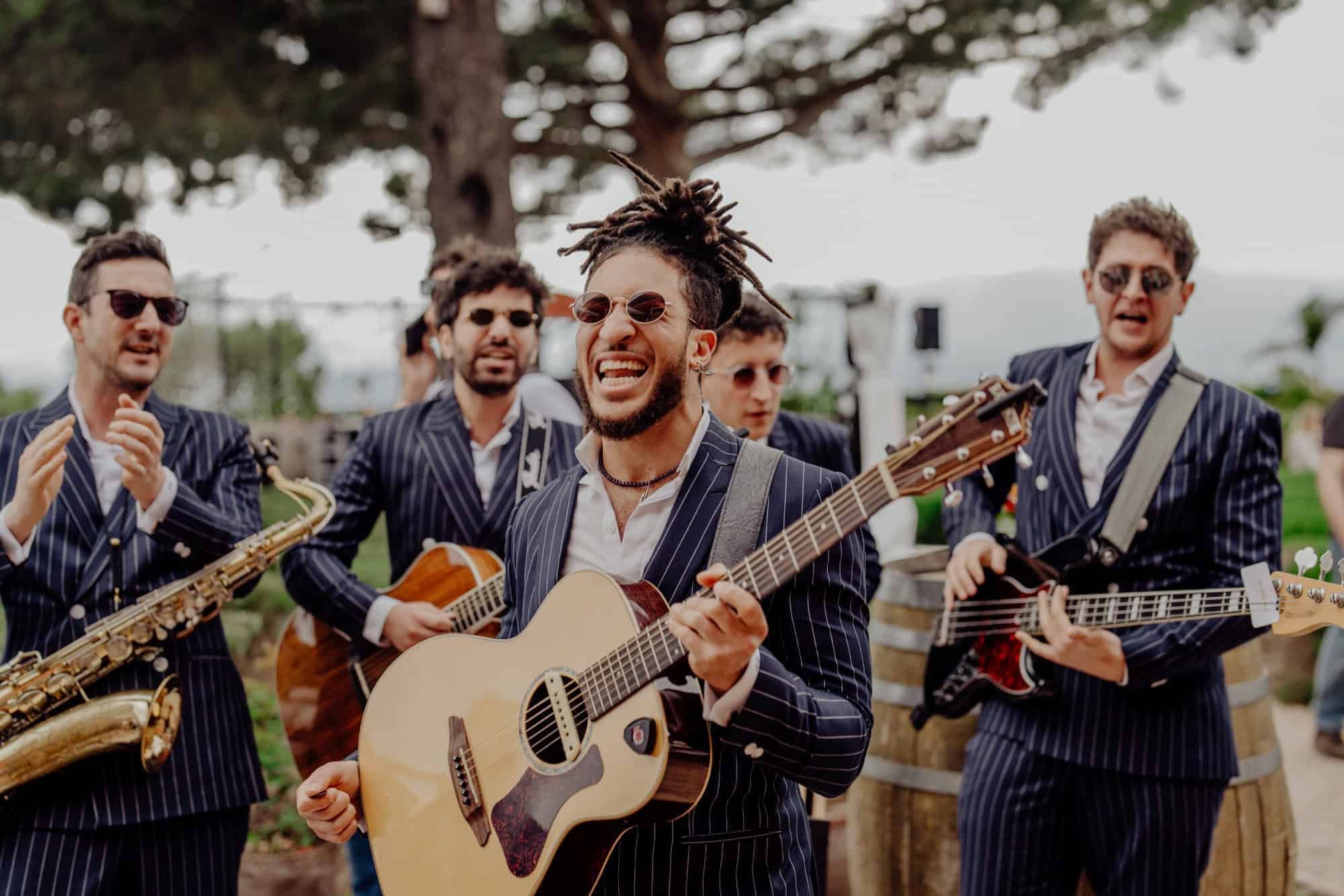 A saxophonist and two guitarists playing outdoors under pine trees