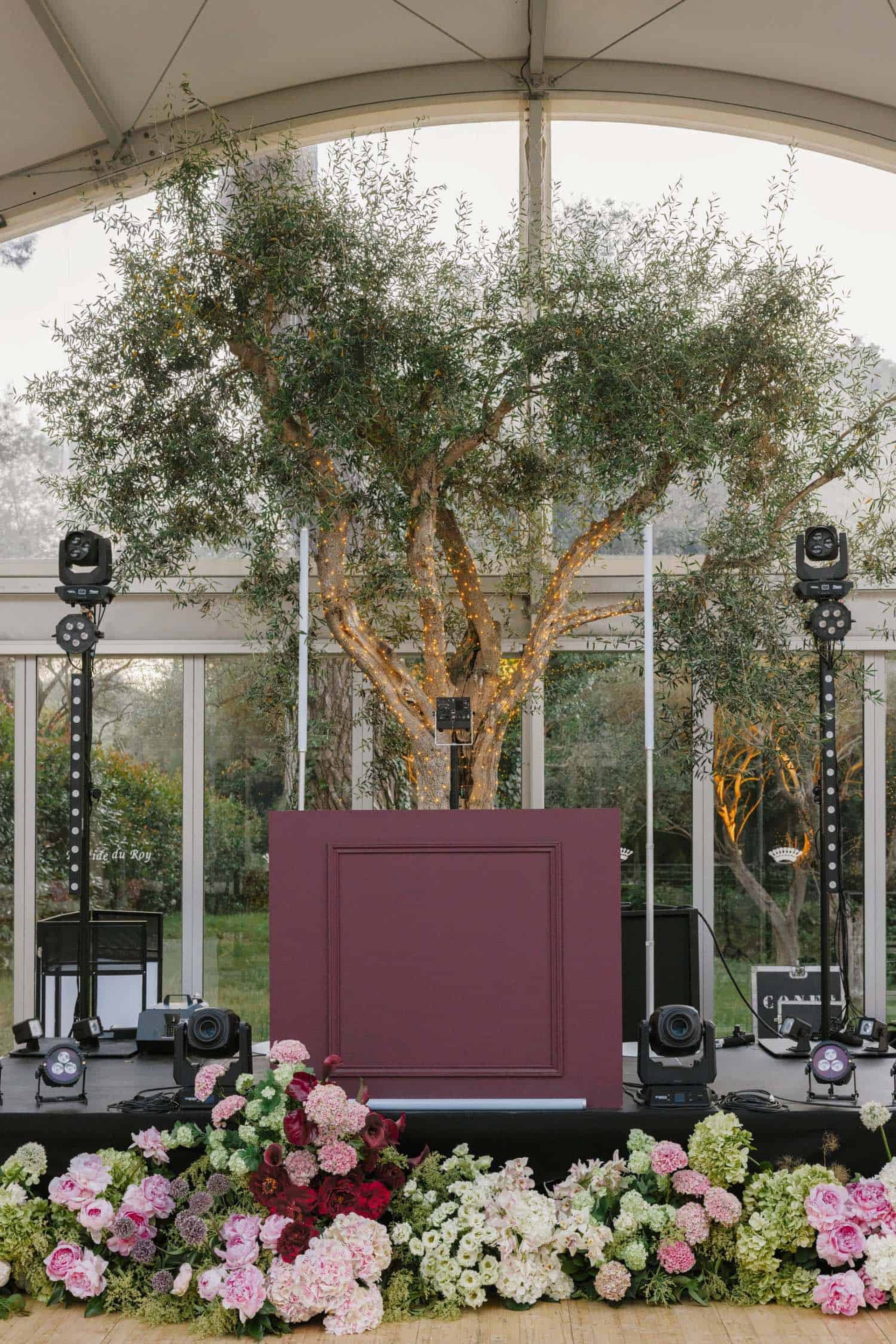 A DJ booth dressed with flowers beneath an olive tree, in a marquee