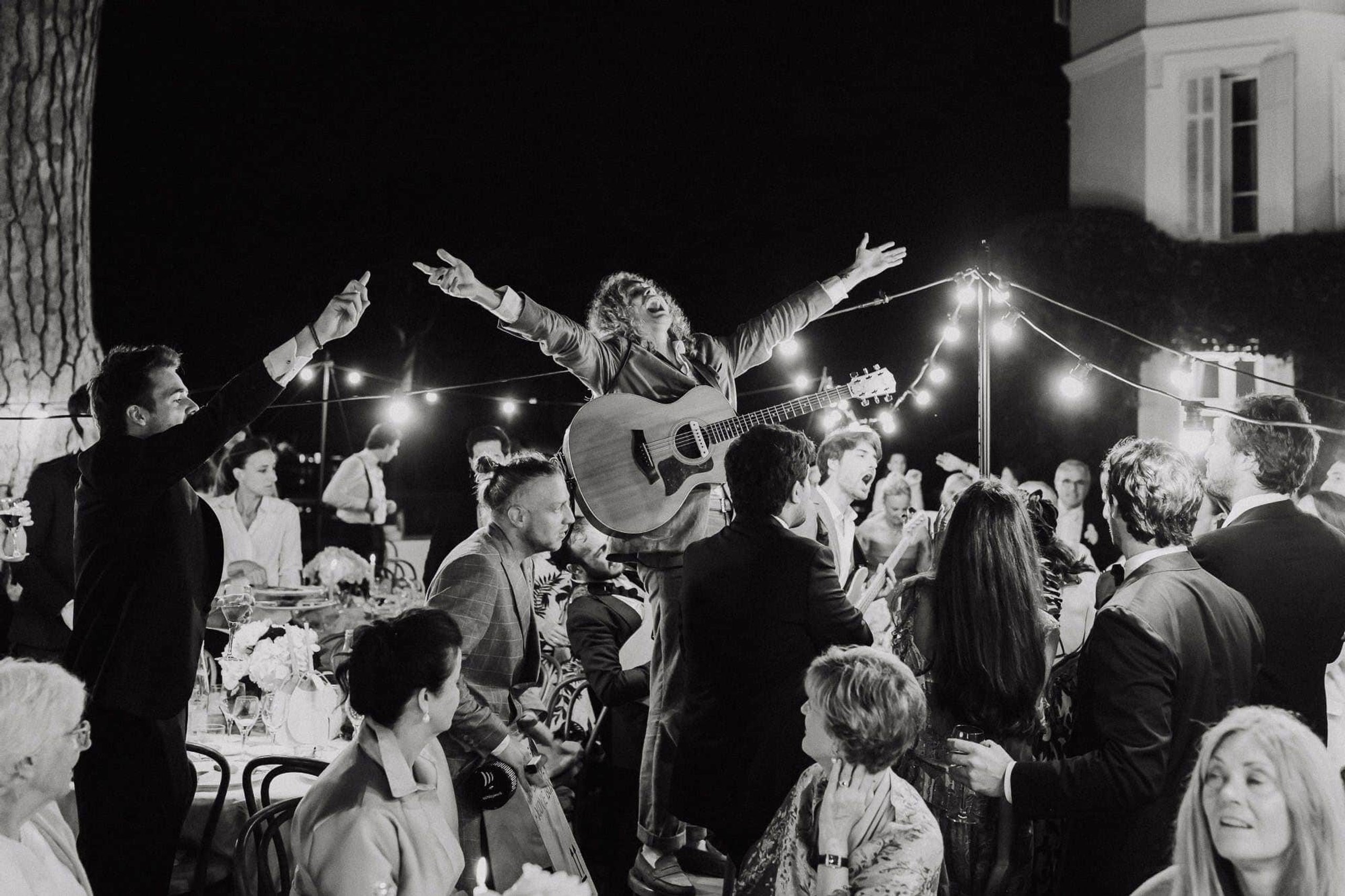 A strolling band playing among guests at an evening dinner under festoon lights