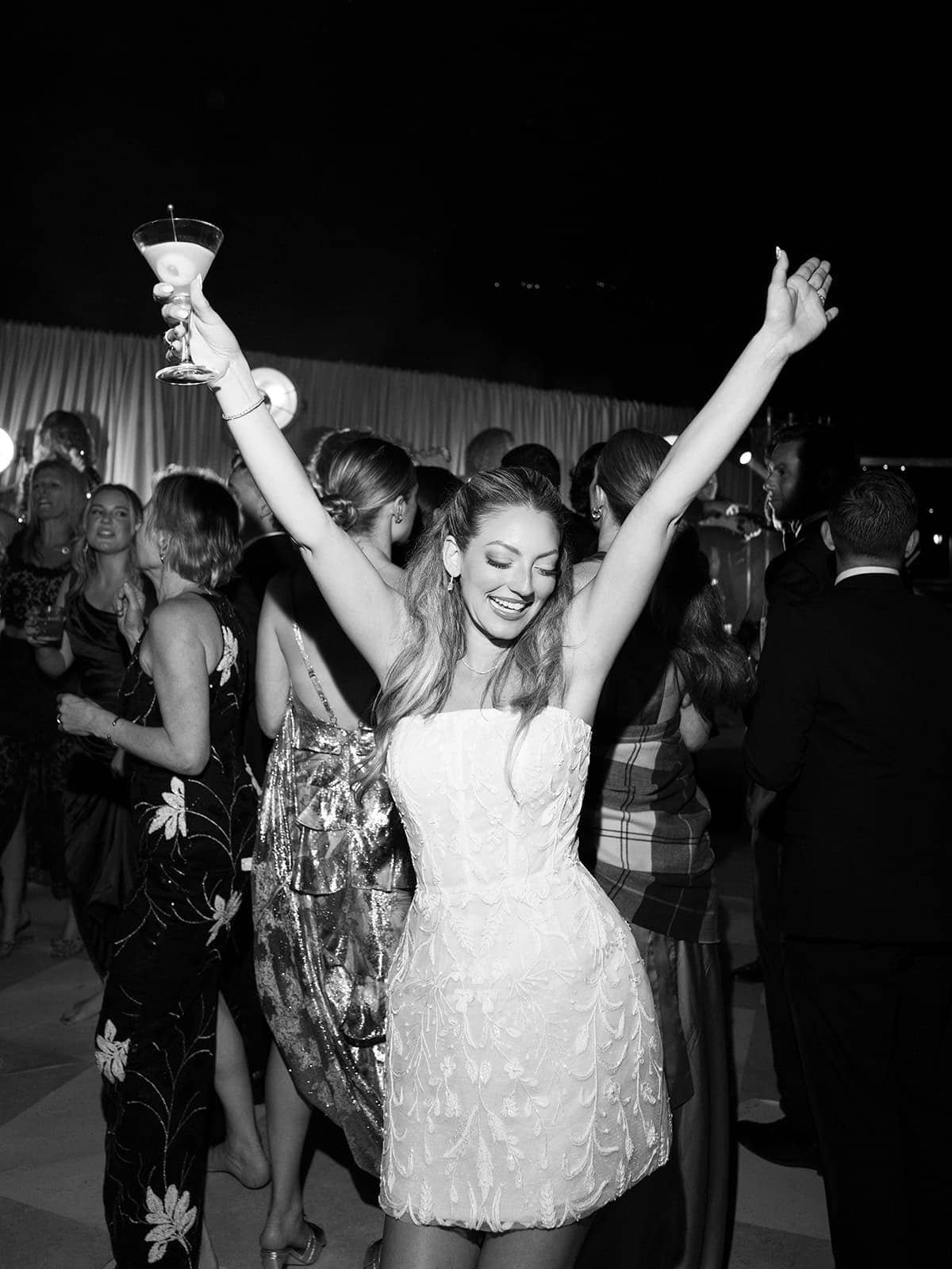 A bride dancing with her arms raised among guests, in black and white