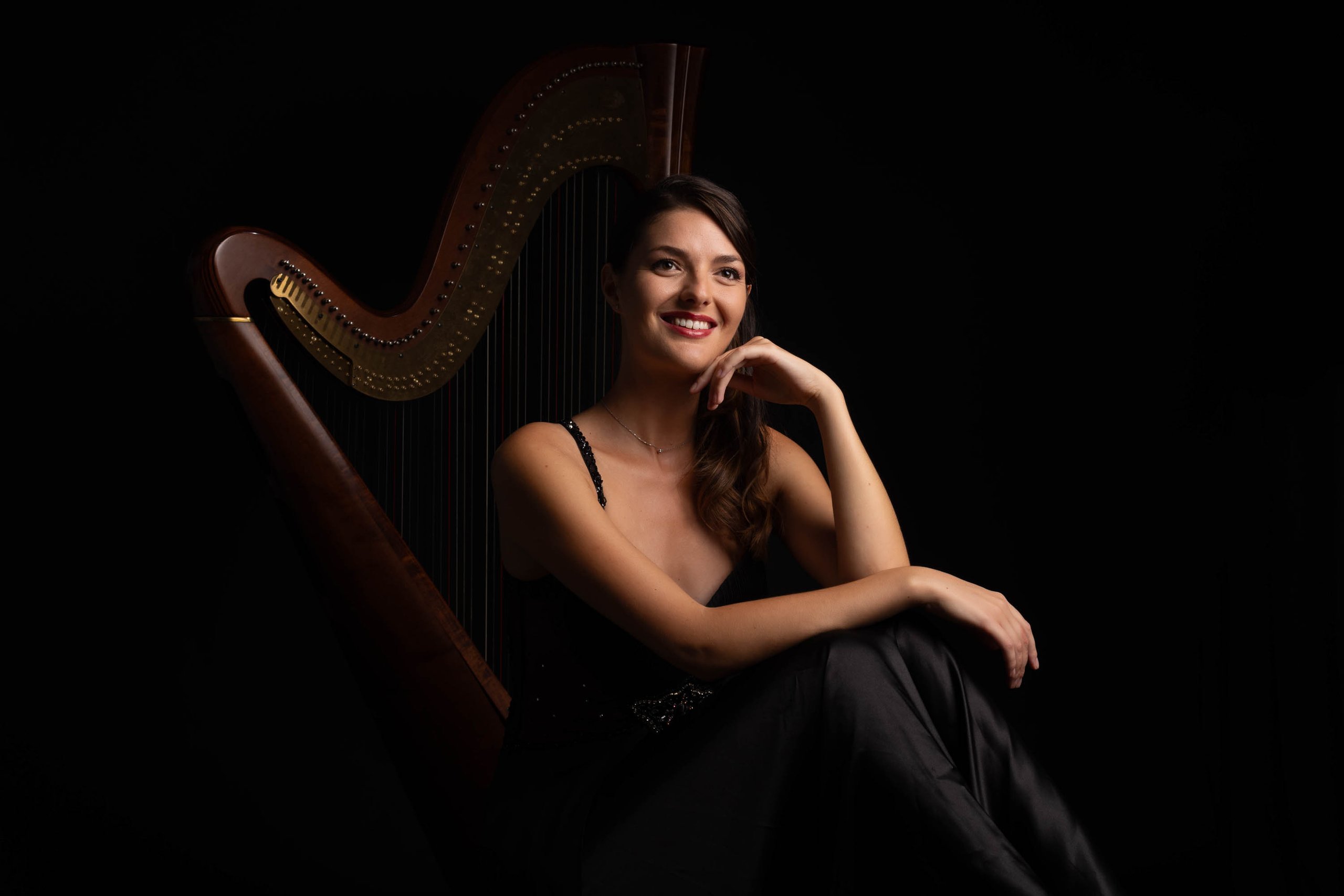 A harpist seated beside her harp against a dark background