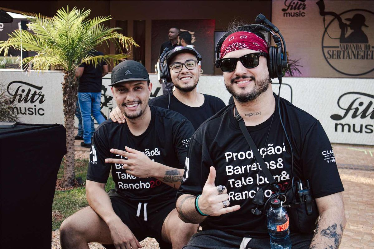 Três integrantes da equipe posando com headsets, palmeiras e o painel Filtr Music ao fundo