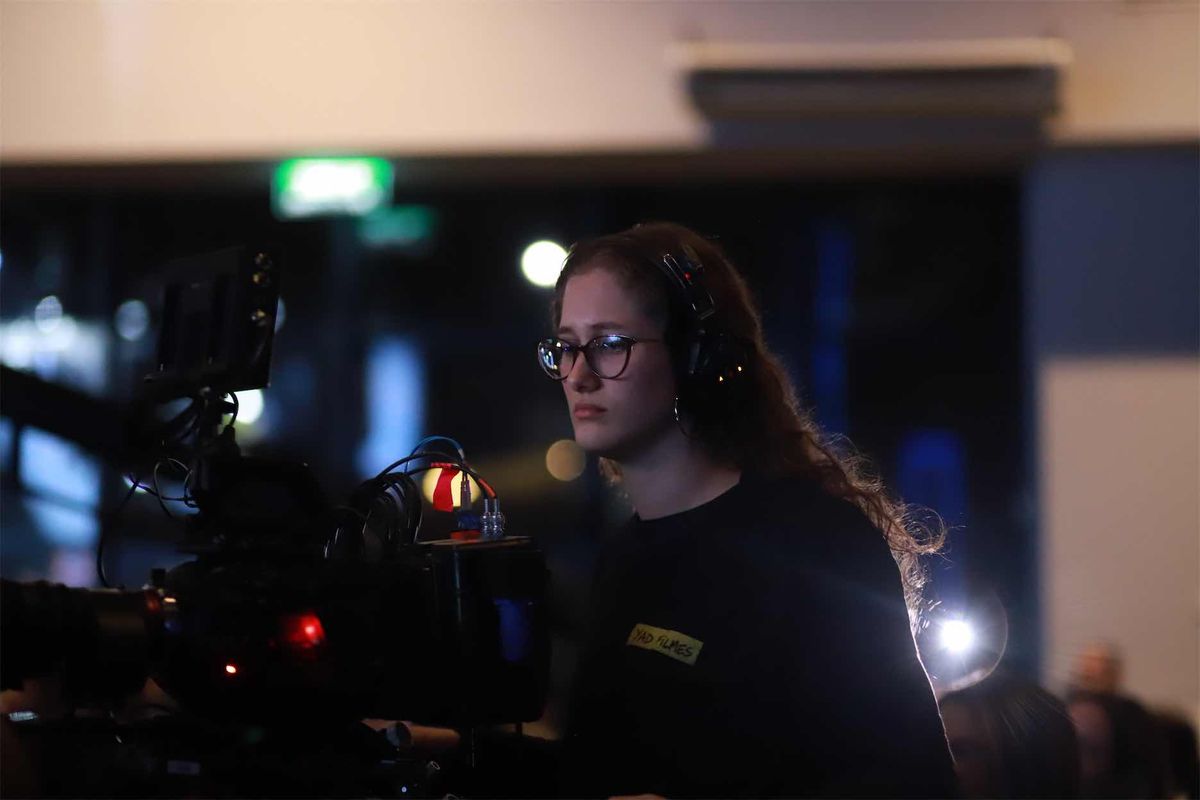 Operadora de headset e óculos atrás da câmera, com etiqueta YAD Filmes na camiseta