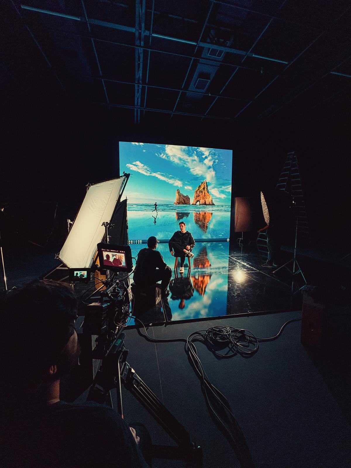 Vista geral do estúdio durante a gravação, com o painel de LED exibindo uma paisagem de praia com formação rochosa, entrevistado sentado ao centro e equipamentos de iluminação nas laterais.