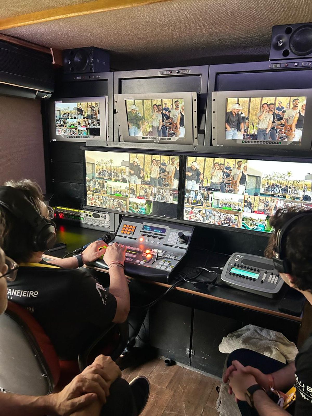 Dois operadores de headset dentro da unidade móvel, diante da mesa de corte e dos monitores de multiview mostrando o palco
