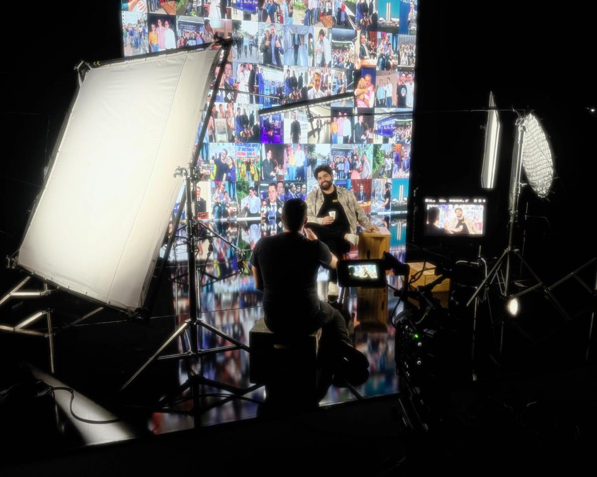 Entrevistado sentado diante do painel de LED exibindo um mosaico de fotos, cercado por refletores e câmera em primeiro plano.