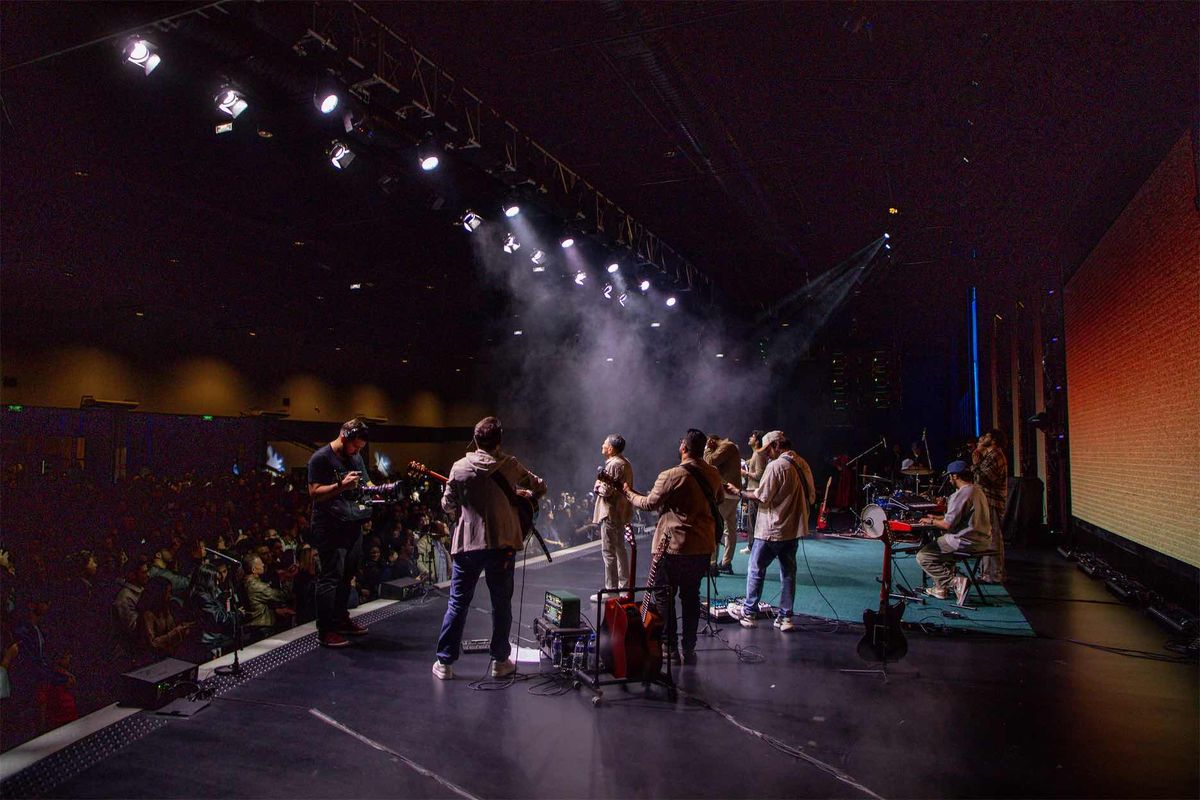 Palco visto da lateral durante a gravação: músicos em cena, plateia à esquerda e telão iluminado à direita