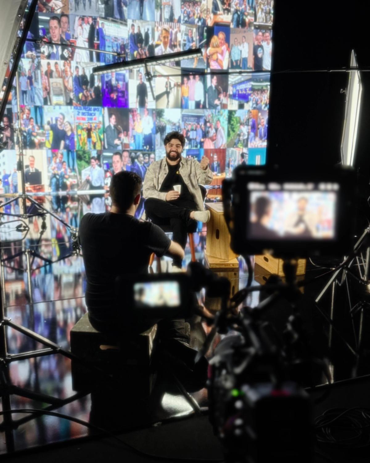 Cinegrafista filmando entrevistado sentado diante do painel de LED com mosaico de fotos, com o enquadramento visível no visor da câmera em primeiro plano.