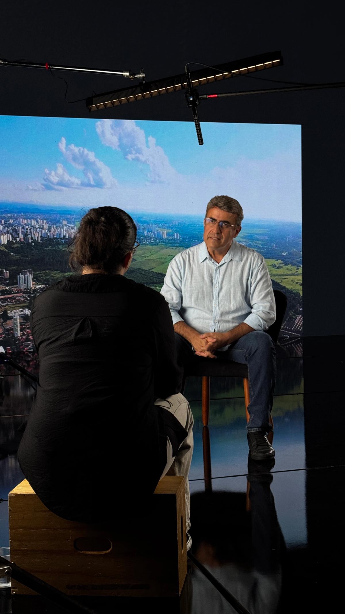 Entrevistado sentado em banqueta diante do painel de LED exibindo uma paisagem aérea de cidade e vegetação, com a entrevistadora de costas em primeiro plano.