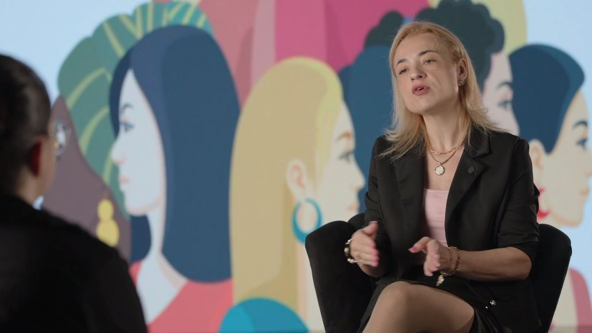 Entrevistada sentada em cadeira preta durante gravação, com painel de LED ao fundo exibindo ilustração colorida de silhuetas femininas.