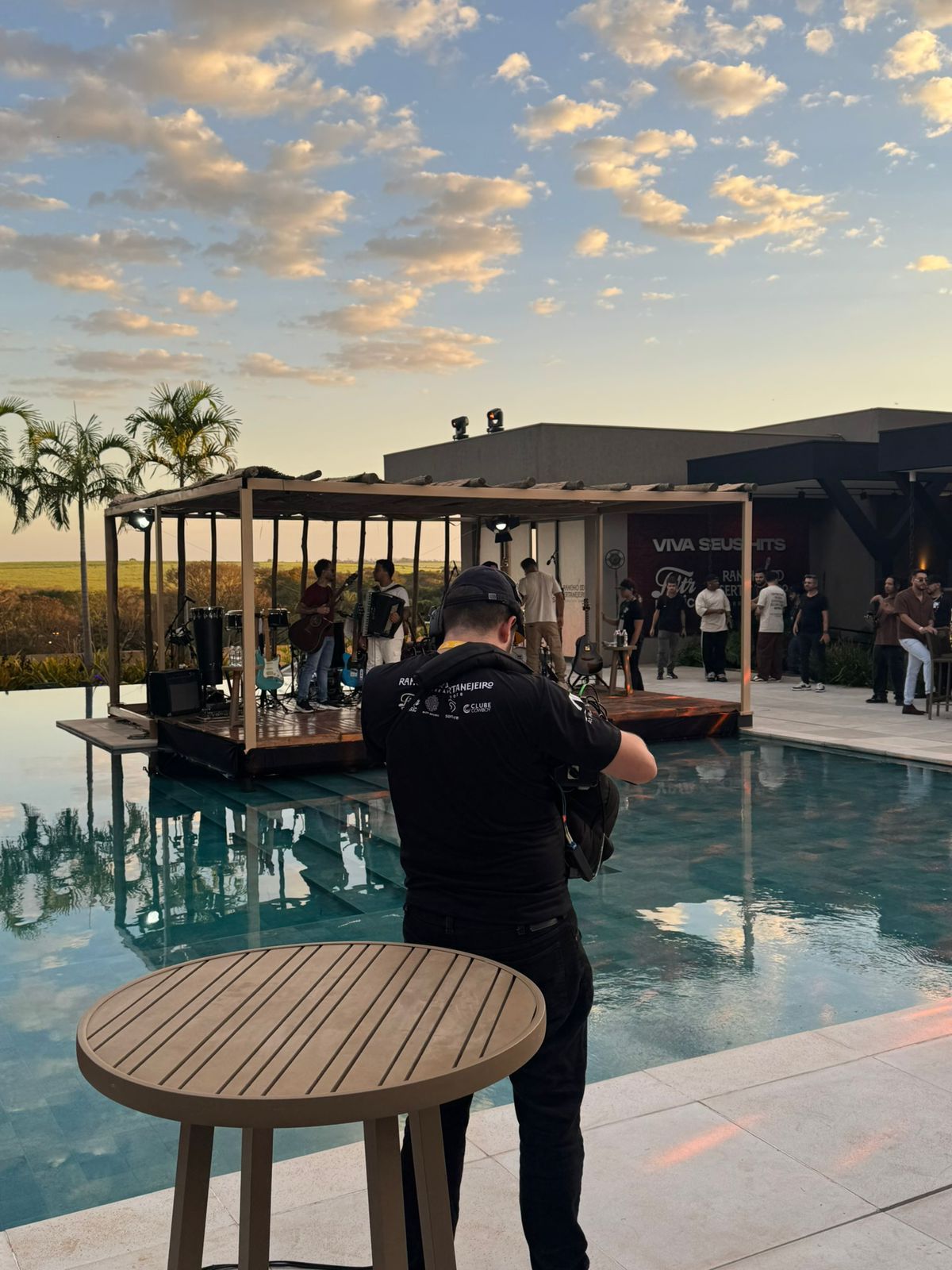 Operador de câmera na beira da piscina ao entardecer, com o palco do Rancho ao fundo