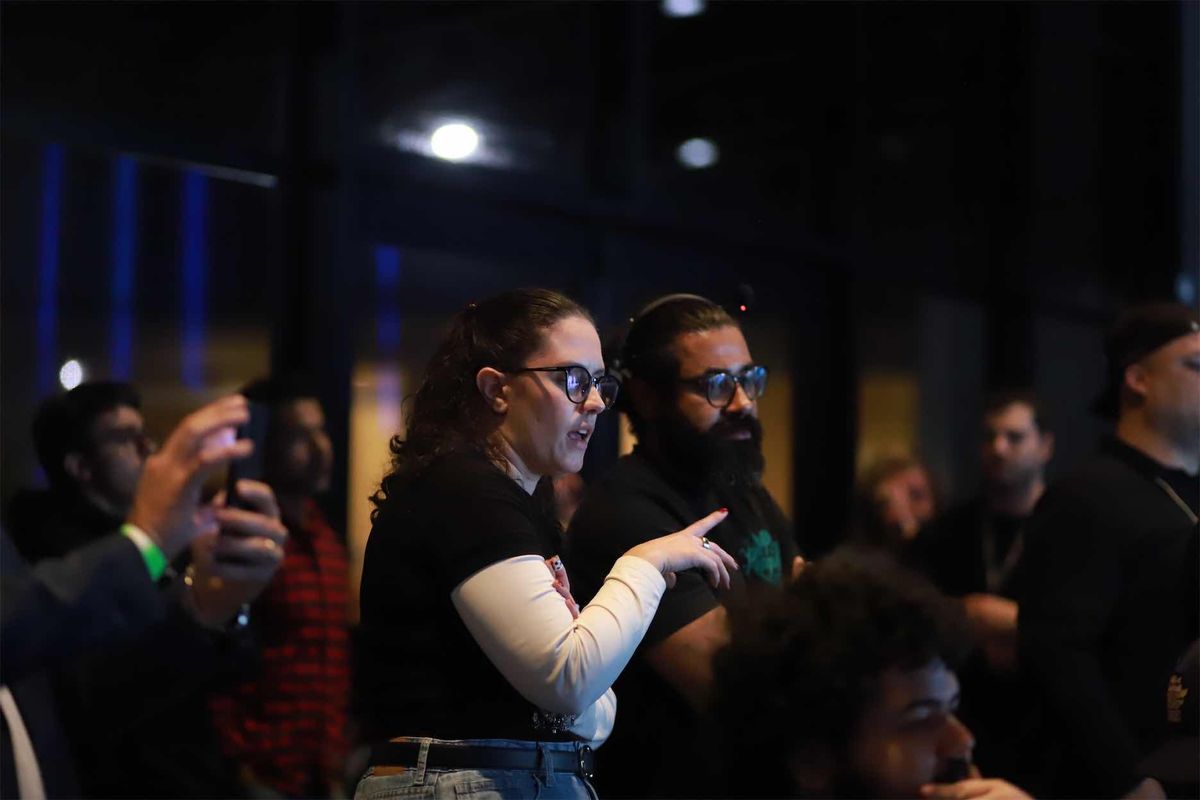 Dois integrantes da equipe de pé no meio do público, um deles apontando uma posição