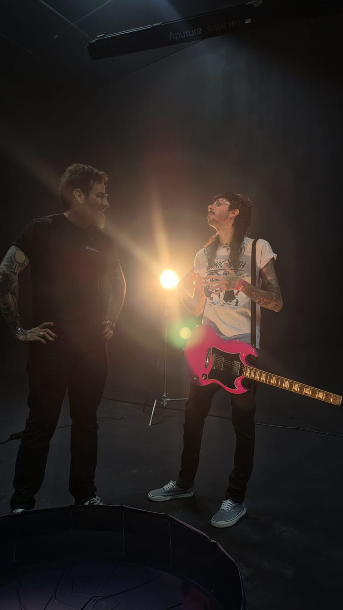 Dois integrantes da equipe rindo entre um take e outro, com a guitarra rosa e um flare de luz