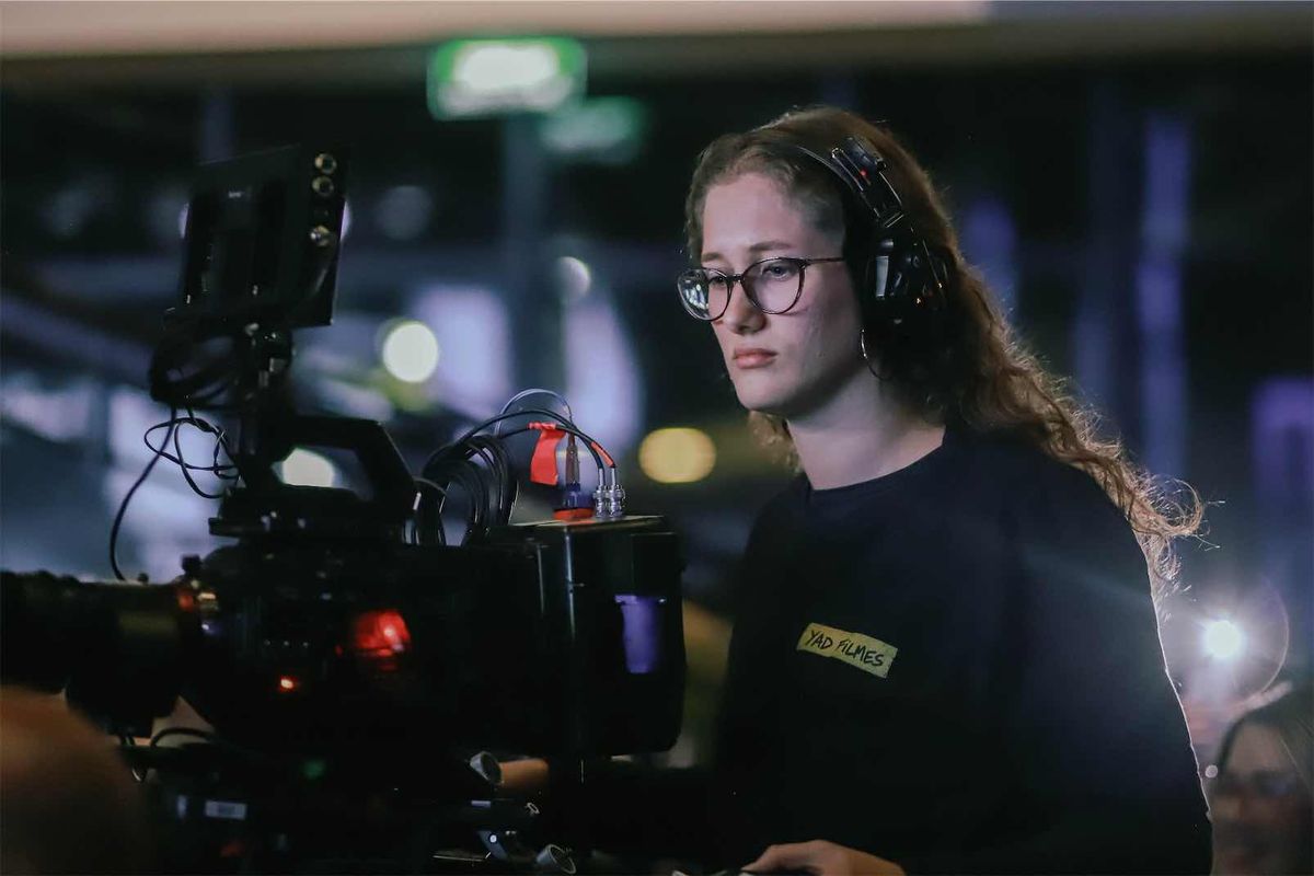 Operadora de cabelo cacheado com headset diante da câmera, iluminada pela luz do set