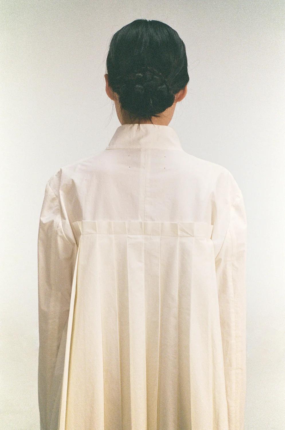 Model from behind in a pleated ivory Unisecon shirt dress, hair in a braided bun