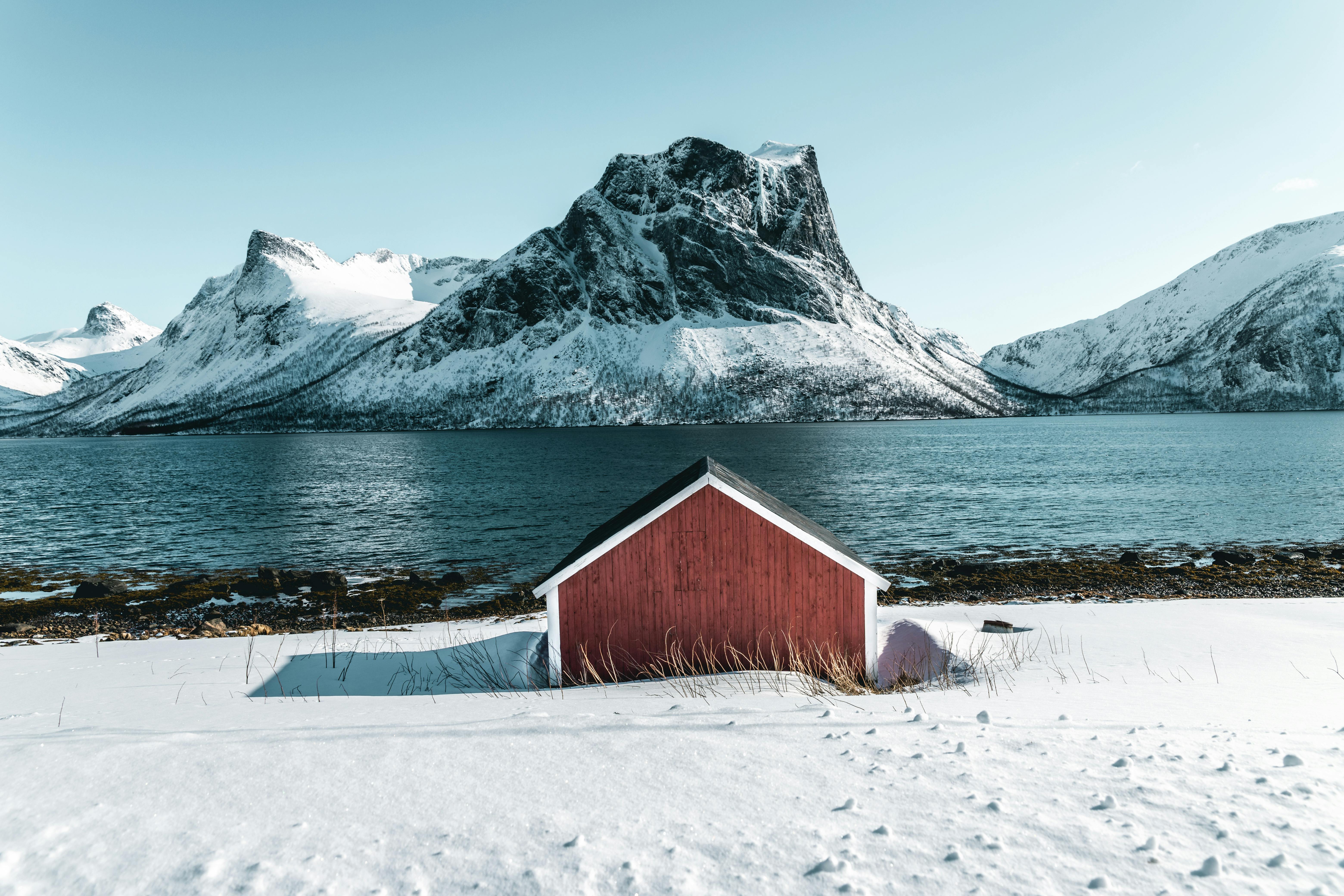 Hytte som ligger ved vannet foran noen store islagte fjell.