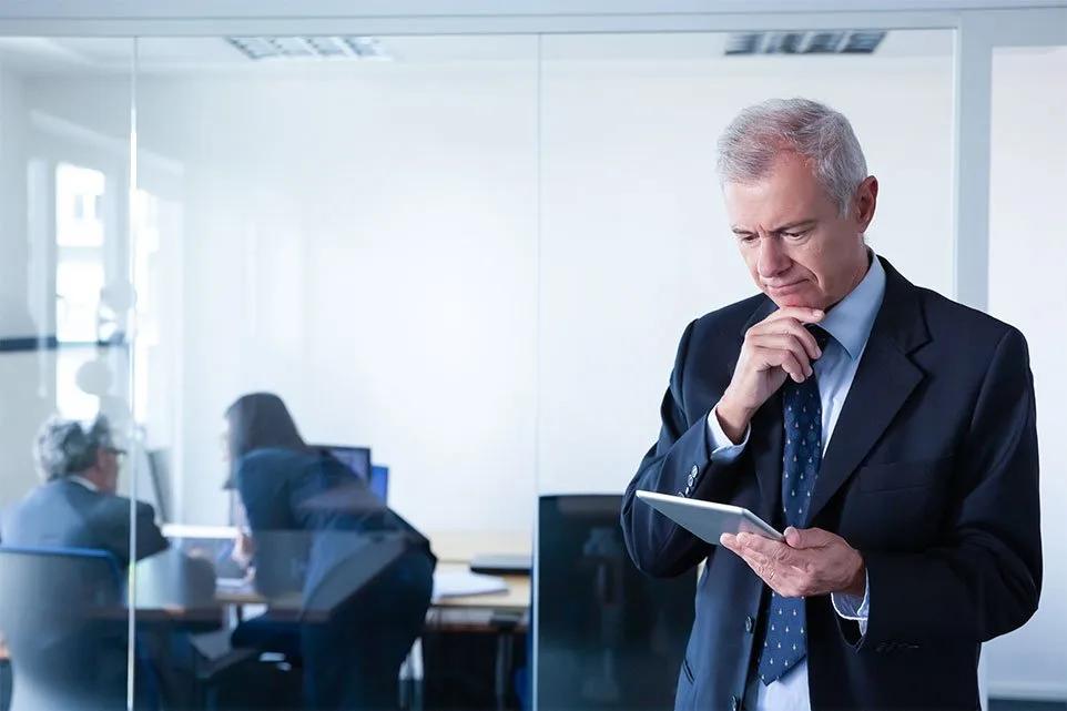 A man in a dark suit stands reading a tablet in a glass-partitioned office