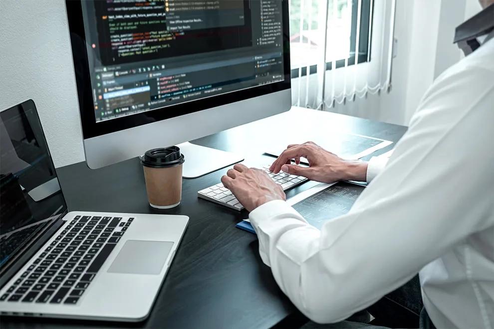 A person at a desk typing at a keyboard in front of a monitor showing syntax-highlighted code