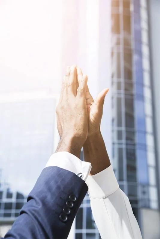 Two raised forearms in suit sleeves meeting in a high-five against a glass office tower